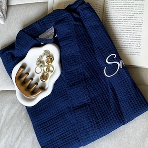 May include: A navy blue waffle-weave robe with the letter "S" embroidered on the front. A white ceramic dish holds gold jewelry and a wooden comb. An open book is visible in the background.