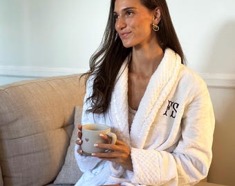 May include: A white, plush bathrobe with the initials "FS" embroidered on the chest. The robe has a shawl collar and is worn with a gray mug with a heart design. A book is on the sofa.