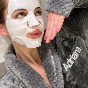 May include: A woman wearing a gray fuzzy robe with the name "Adriana" embroidered on the front. She is wearing a white face mask.