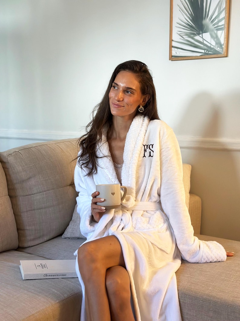 May include: A white, plush robe with the initials "FS" embroidered on the chest. The robe is tied at the waist and worn with a matching belt. A beige mug with a heart design is held in the hand. The person is sitting on a beige couch.