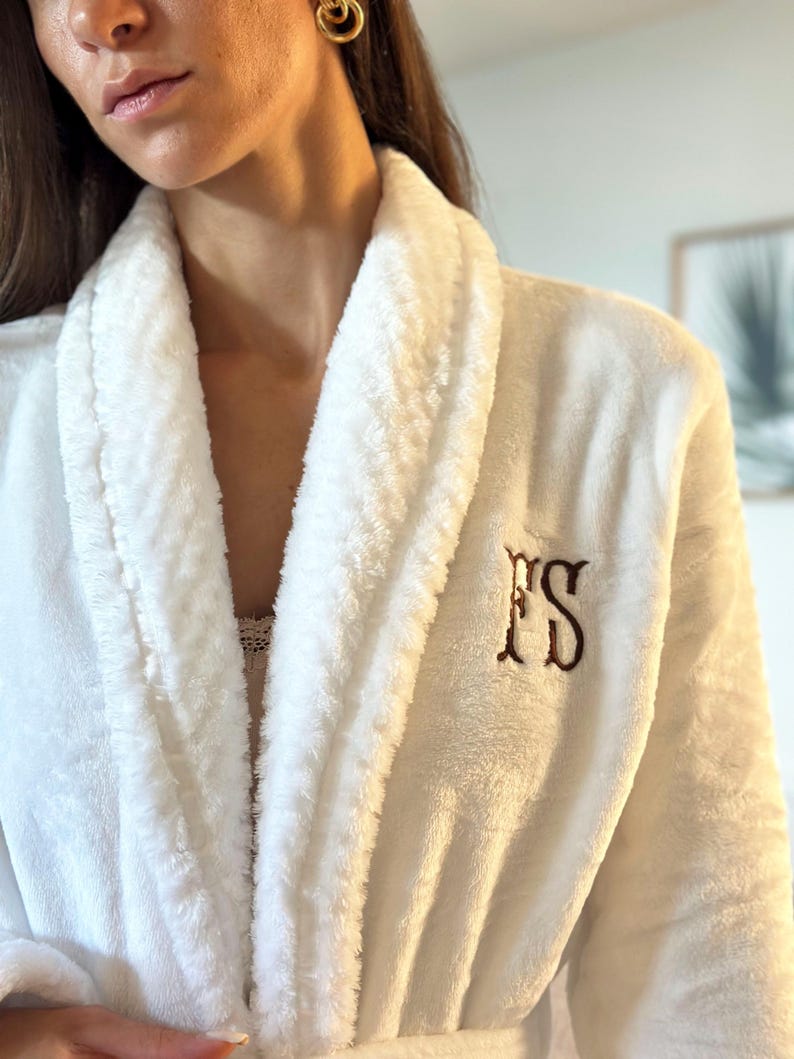 May include: A white plush robe with a shawl collar and a matching belt. The robe features embroidered initials "FS" in a brown color on the chest. The robe is made of a soft, textured material.