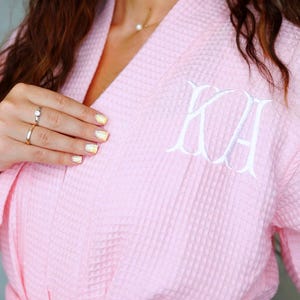 May include: A light pink waffle-knit robe featuring a white embroidered monogram with the letters KA on the left chest. The robe has a wrap-front design with a matching fabric tie belt at the waist and long sleeves, showcasing a textured fabric.