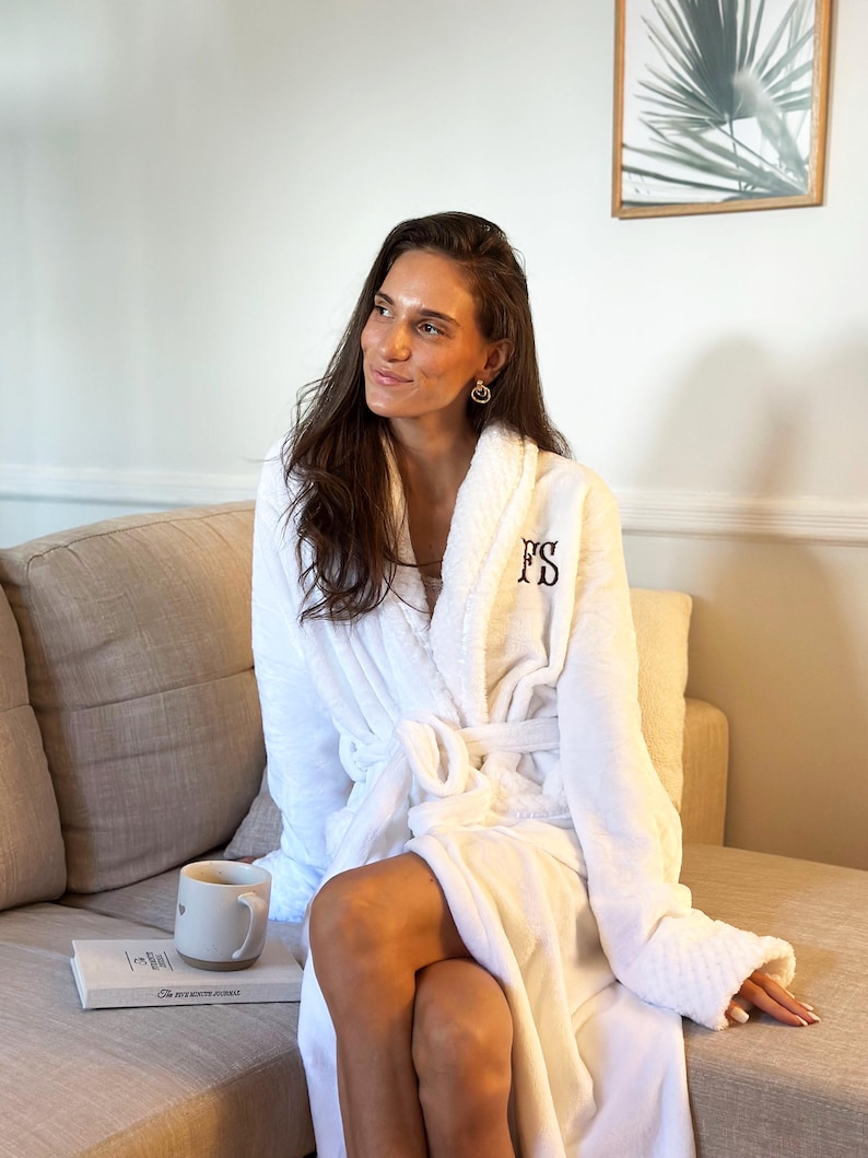 May include: A white, plush bathrobe with the embroidered initials "FS" in black. The robe has a shawl collar, a waist tie, and long sleeves. A coffee mug and a book are on a nearby surface.