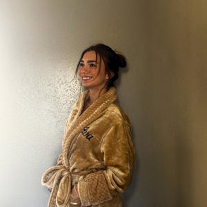 May include: A plush, golden-brown robe with a shawl collar and a matching tie belt. The robe features the embroidered name "Ava" in a cursive font on the chest. The cuffs and collar have a textured design.