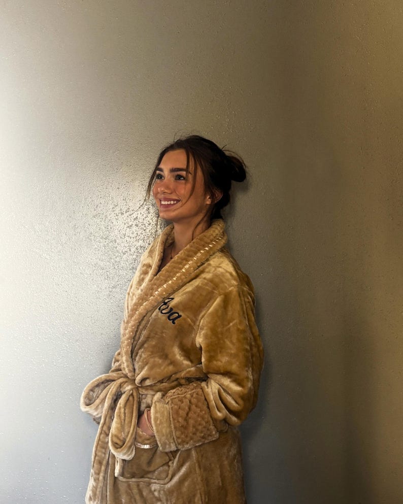 May include: A plush, golden-brown robe with a shawl collar and a matching tie belt. The robe features the embroidered name "Ava" in a cursive font on the chest. The cuffs and collar have a textured design.