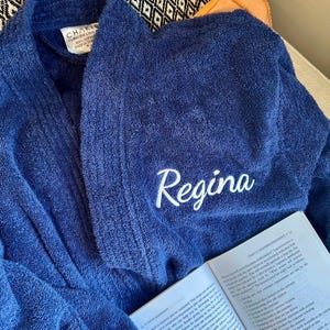 May include: A navy blue terry cloth bathrobe with the name "Regina" embroidered in white. The robe is folded over an open book. The label reads "CHAKIR TURKISH LINE".