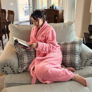 May include: A person wearing a pink plush robe with the word "Victoria" embroidered on the chest, sitting on a gray sofa and reading a book. The sofa has two plaid pillows and is in a well-lit living room.