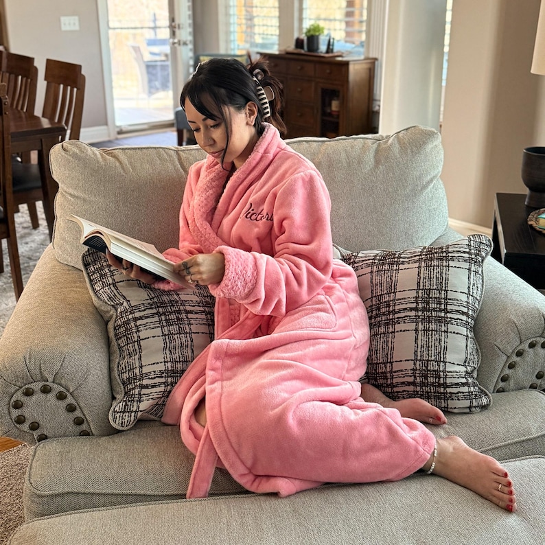 May include: A person wearing a pink plush robe with the word "Victoria" embroidered on the chest, sitting on a gray sofa and reading a book. The sofa has two plaid pillows and is in a well-lit living room.