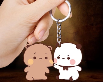 Puede incluir: Un llavero con dos personajes de osos de dibujos animados. Uno es marrón con una mejilla amarilla, el otro blanco con detalles marrones. El llavero tiene un anillo y una cadena plateados.