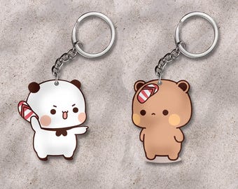 May include: Two keychains featuring cartoon characters. One is a white panda holding a red and white striped flip-flop. The other is a brown bear with a red and white striped bandage on its head. Both have silver key rings.