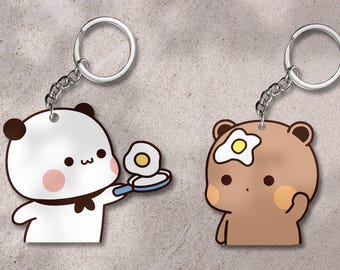 May include: Two keychains featuring cartoon bear and panda characters. One keychain shows a panda holding a frying pan with a fried egg. The other keychain shows a bear with a fried egg on its head. Both keychains have silver key rings.