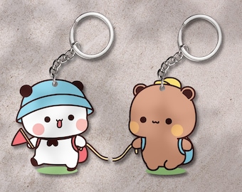 May include: Two keychains featuring cartoon bear characters. One bear is white with a blue hat and pink backpack, holding a red flag. The other is brown with a yellow hat and blue backpack. Both are connected by a yellow string.