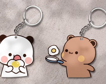 May include: Two cartoon character keychains. One is a white panda holding a fried egg, the other is a brown bear holding a frying pan with a fried egg. Both keychains have silver key rings and chains.