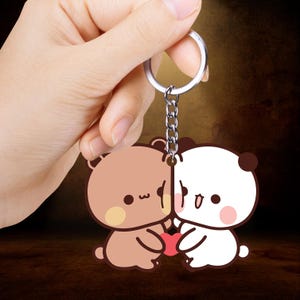 Puede incluir: Un llavero con dos osos de dibujos animados, uno marrón y otro blanco, sosteniendo un corazón rojo. Los osos tienen ojos negros y están conectados por una cadena y un llavero plateados. El fondo es marrón borroso.