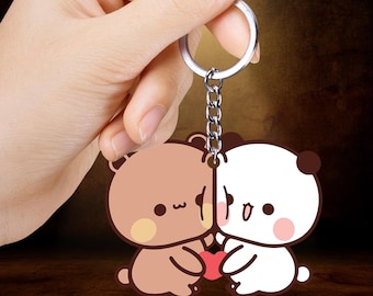 May include: A keychain featuring two cartoon bears, one brown and one white, holding a red heart. The bears have black eyes and are connected by a silver chain and keyring. The background is a blurred brown.
