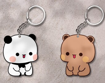 Puede incluir: Dos llaveros con diseños de oso y panda de dibujos animados. El panda es blanco con detalles negros y una pajarita. El oso es marrón con una lengua rosa. Ambos tienen llaveros y cadenas plateadas.