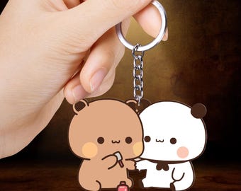 May include: A keychain featuring two cartoon bears. One bear is brown and holding a small brush and a pink bottle. The other bear is white with black accents. The keychain has a silver chain and ring.