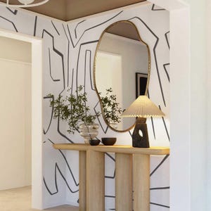 May include: An entryway with a light wood console table, a gold-framed mirror, and a lamp with a pleated shade. The wall is decorated with a black and white abstract pattern, and a vase with greenery sits on the table.