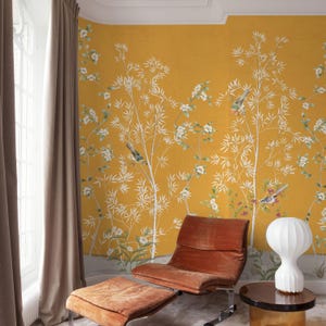 May include: A room with mustard yellow wallpaper featuring a white bamboo design with birds and flowers. A brown chair and ottoman sit in front of the wallpaper. A white lamp and a small table are also in the room. Brown curtains frame a window.