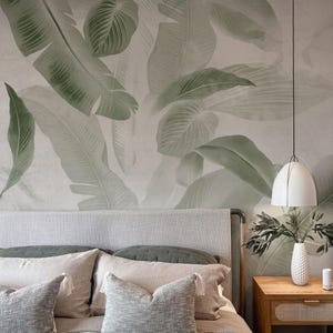 May include: A bedroom interior with a bed, featuring a grey headboard and pillows. The wall is decorated with a large green and white banana leaf wallpaper. A wooden bedside table with a white vase and a pendant light complete the scene.