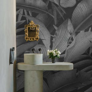 May include: A modern bathroom with a gray and white tropical leaf wallpaper. A gold framed mirror hangs above a concrete sink and countertop. A small vase of white flowers sits on the countertop.