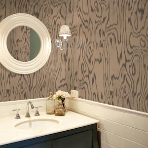 May include: A bathroom vanity with a white marble countertop, a chrome faucet, and a white circular mirror. The wall features a brown and gray wood grain pattern wallpaper, a chrome wall sconce, and a white electrical outlet above white tiles.