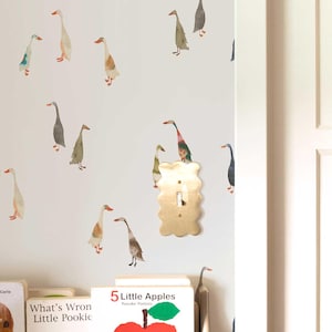 May include: A white wall adorned with watercolor goose decals in various colors. A gold-colored light switch is mounted on the wall. Children's books, including "5 Little Apples," are visible on a shelf below.