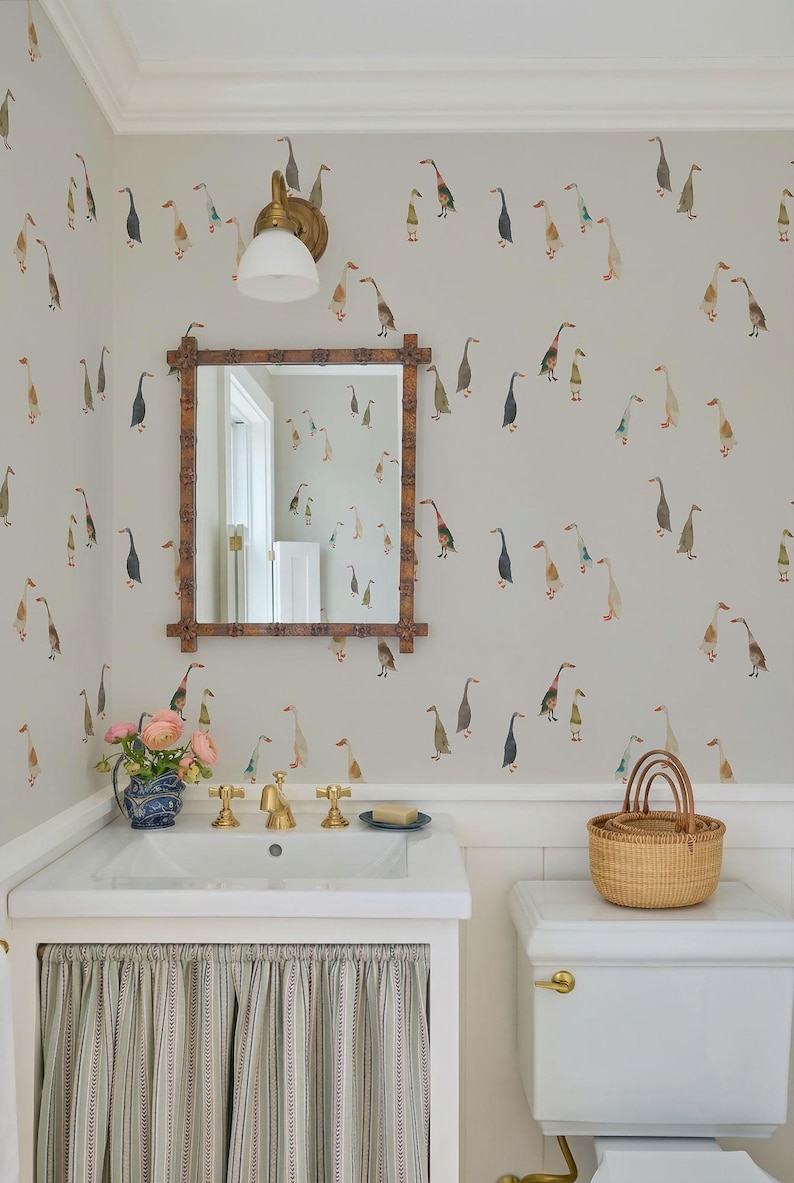 a bathroom with a white sink, a mirror, a toilet, and a basket on the toilet tank. The walls are decorated with a pattern of ducks and geese, and there is a vase of flowers on the sink.