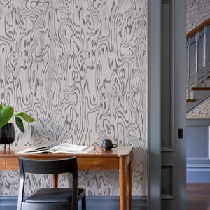 May include: A room featuring wallpaper with a gray and white wood grain pattern, a wooden desk with an open book and a black mug, and a black chair. A doorway with blue trim leads to a staircase with a wooden handrail and matching wallpaper.