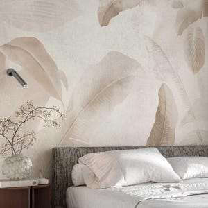May include: A bedroom scene featuring a large-scale wallpaper with a botanical leaf design in shades of beige and cream. The bed has white linens and pillows, with a small side table holding a vase and books. A modern wall lamp is visible.