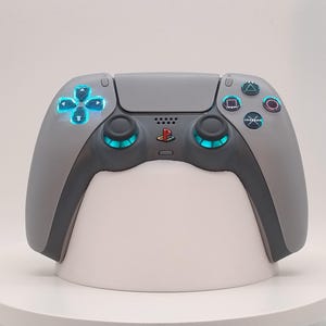 May include: A grey and black games controller with illuminated blue buttons and directional pad. The controller features a PlayStation logo and is displayed on a white stand. The controller's design is modern and sleek.