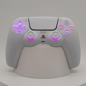 May include: A white PlayStation 5 controller with pink and multi-coloured illuminated buttons. The controller is on a white stand, highlighting its sleek design and ergonomic shape. The PlayStation logo is visible.