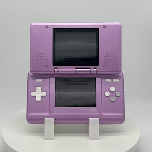 May include: A lavender Nintendo DS handheld gaming console, open to display two screens. The bottom screen features a directional pad and buttons, while the top screen is angled upward. The console is resting on a white stand.