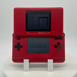 May include: A red Nintendo DS handheld game console, open to display two screens. The top screen shows a clock and calendar, while the bottom screen displays game information. The console has a directional pad, buttons, and a stylus slot.