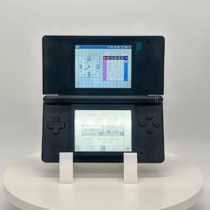May include: Black Nintendo DS handheld game console, open to display two screens. The top screen shows a clock and calendar, while the bottom screen displays text. The console is on a white stand.