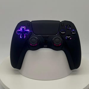 Könnte beinhalten: Schwarzer Gaming-Controller mit lila und rosa beleuchteten Akzenten. Der Controller verfügt über ein Steuerkreuz, Aktionstasten und Analog-Sticks. Er liegt auf einem weißen Ständer.