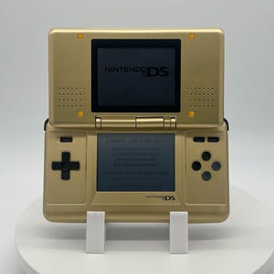 May include: Gold-colored Nintendo DS handheld game console, open and displayed on a white stand. The top screen displays the Nintendo DS logo. The bottom screen shows text. The console has a directional pad, buttons, and a stylus slot.