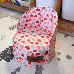 Puede incluir: Una silla pequeña, redonda y tapizada con una base de tela blanca cubierta por un patrón repetitivo de corazones rojos y rosas. La silla tiene un respaldo curvo y un parche rectangular marrón de aspecto de cuero en el frente con el texto UMAY.