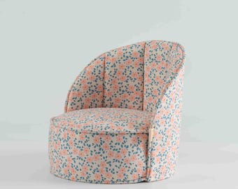 May include: A small, rounded armchair upholstered in a cream-colored fabric featuring a repeating pattern of soft pink flowers and small blue leaves. The chair has a curved backrest and a low, cylindrical seat with a small fabric tag on the side.