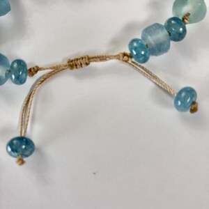 Op de afbeelding: Close-up van een armband met blauwe en aquakleurige kralen. De kralen zijn geregen op een beige koord, met een schuifknoopsluiting. De kralen vari&euml;ren in vorm en grootte, wat een unieke, handgemaakte uitstraling cre&euml;ert.