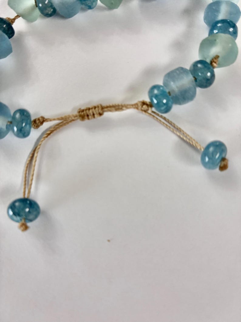 Op de afbeelding: Close-up van een armband met blauwe en aquakleurige kralen. De kralen zijn geregen op een beige koord, met een schuifknoopsluiting. De kralen vari&euml;ren in vorm en grootte, wat een unieke, handgemaakte uitstraling cre&euml;ert.