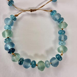 Peut inclure: Bracelet réglable composé de perles de verre bleues et vertes. Les perles, de tailles et formes variées, sont enfilées sur un cordon beige. Le bracelet est doté d'une fermeture coulissante réglable. Un bijou simple et élégant.
