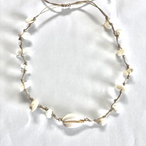 Könnte beinhalten: Eine naturfarbene Halskette mit kleinen weißen Muscheln und Muschelsplittern, die auf einer beigefarbenen Kordel aufgereiht sind. Die Halskette ist in der Länge verstellbar und hat einen größeren Muschelanhänger.