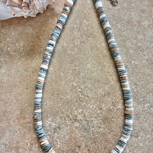 May include: A necklace composed of small, flat, circular beads in shades of slate blue, white, and tan. The beads are strung in a repeating pattern, and the necklace features a silver-toned metal lobster claw clasp with an adjustable chain extension.