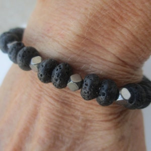 May include: A black lava rock bracelet with silver-colored, hexagonal beads. The bracelet is made of porous, dark gray beads strung together. The bracelet is shown on a person's wrist.