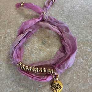 May include: A handmade bracelet featuring a tie-on design with a pink and purple silk ribbon. The bracelet includes a strand of gold beads and a gold charm with an eye symbol.