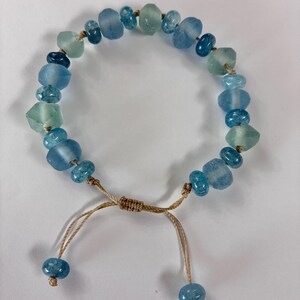 Op de afbeelding: Verstelbare armband met blauwe en groene glazen kralen. De kralen, in verschillende vormen en maten, zijn geregen op een beige koord. De armband heeft een schuifknoopsluiting met twee blauwe kralen aan de uiteinden.