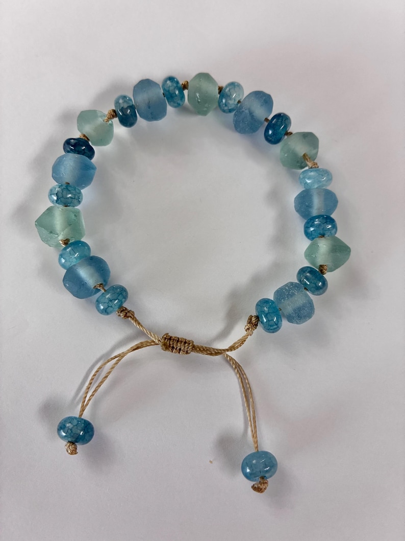 Op de afbeelding: Verstelbare armband met blauwe en groene glazen kralen. De kralen, in verschillende vormen en maten, zijn geregen op een beige koord. De armband heeft een schuifknoopsluiting met twee blauwe kralen aan de uiteinden.