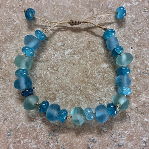 Op de afbeelding: Handgemaakte armband met blauwe en turquoise glazen kralen. De kralen zijn geregen op een beige koord, met een verstelbare schuifknoopsluiting. De armband heeft kralen in verschillende maten en vormen.