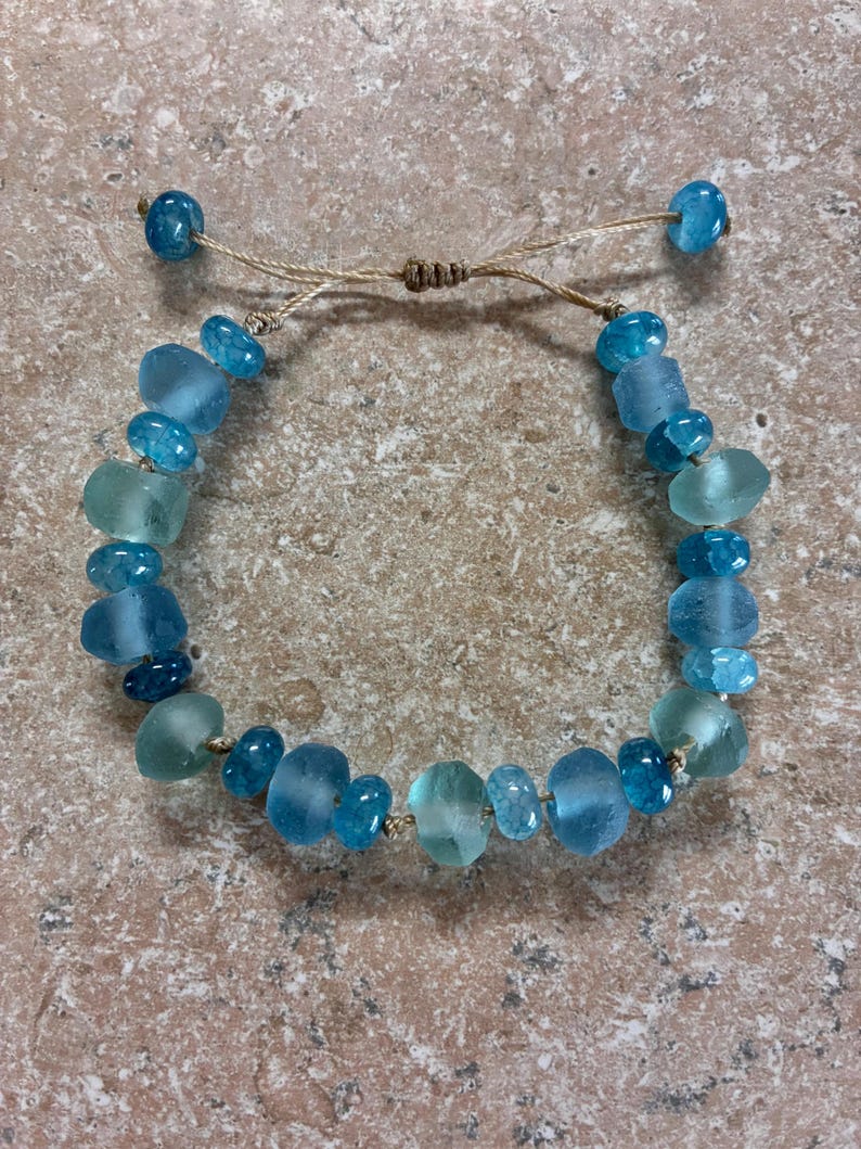 Op de afbeelding: Handgemaakte armband met blauwe en turquoise glazen kralen. De kralen zijn geregen op een beige koord, met een verstelbare schuifknoopsluiting. De armband heeft kralen in verschillende maten en vormen.
